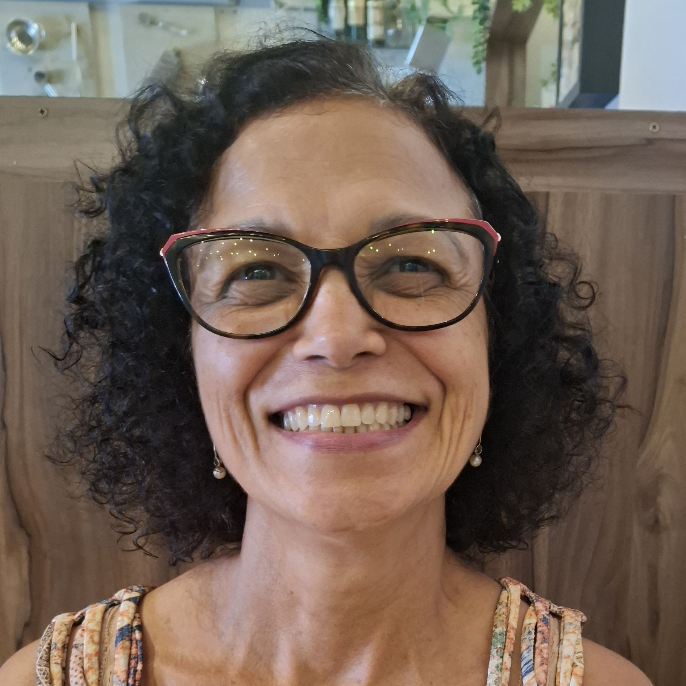 ImagemIlda é uma mulher adulta, cisgênero, morena, olhos castanhos, cabelos escuros, acima dos ombros e cacheados. Na foto, está sorrindo, usa óculos, brincos de pérola e vestido colorido de tons claros, com alças tipo regata