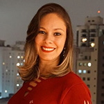 Amanda Ramalho é uma mulher adulta de cabelos escovados castanho escuros com as pontas loiras na altura dos ombros. Na foto está sorrindo e usa uma blusa vermelha