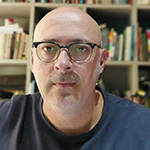 Carlos Gonçalves é um homem adulto cisgênero, branco, com olhos verdes escuros, calvo, usa barba por fazer e óculos de grau com armação retangular preta. Na foto, usa uma camiseta na cor azul marinho. E no fundo, uma estante de madeira com separações e livros diversos