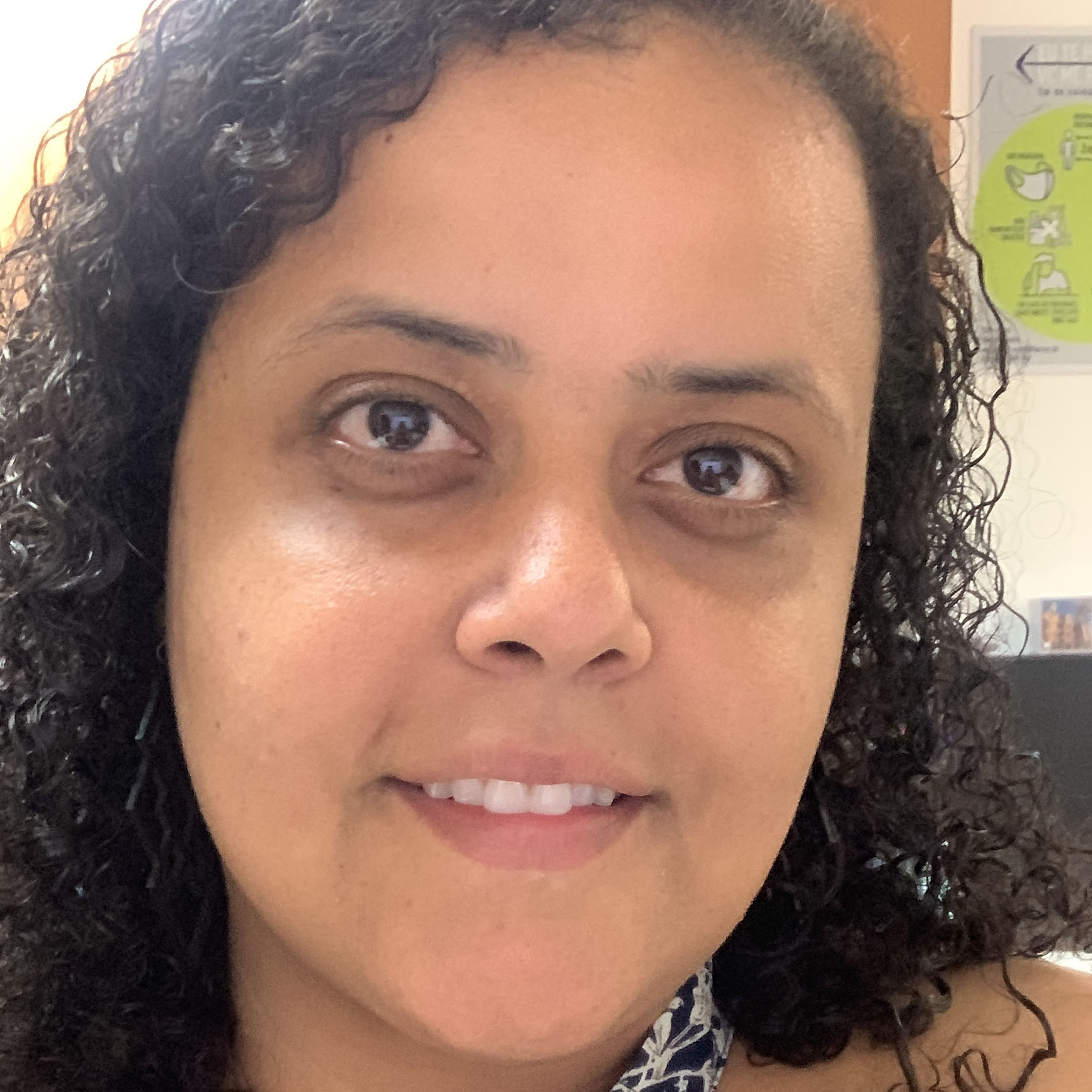 Fotografia de Carolina, uma mulher negra de pele parda, com cabelos castanhos, crespos e encaracolados na altura dos ombros. Tem olhos castanhos e usa camiseta preta com flores brancas. Ela aparece do busto para cima. Está em um ambiente interno, de escritório