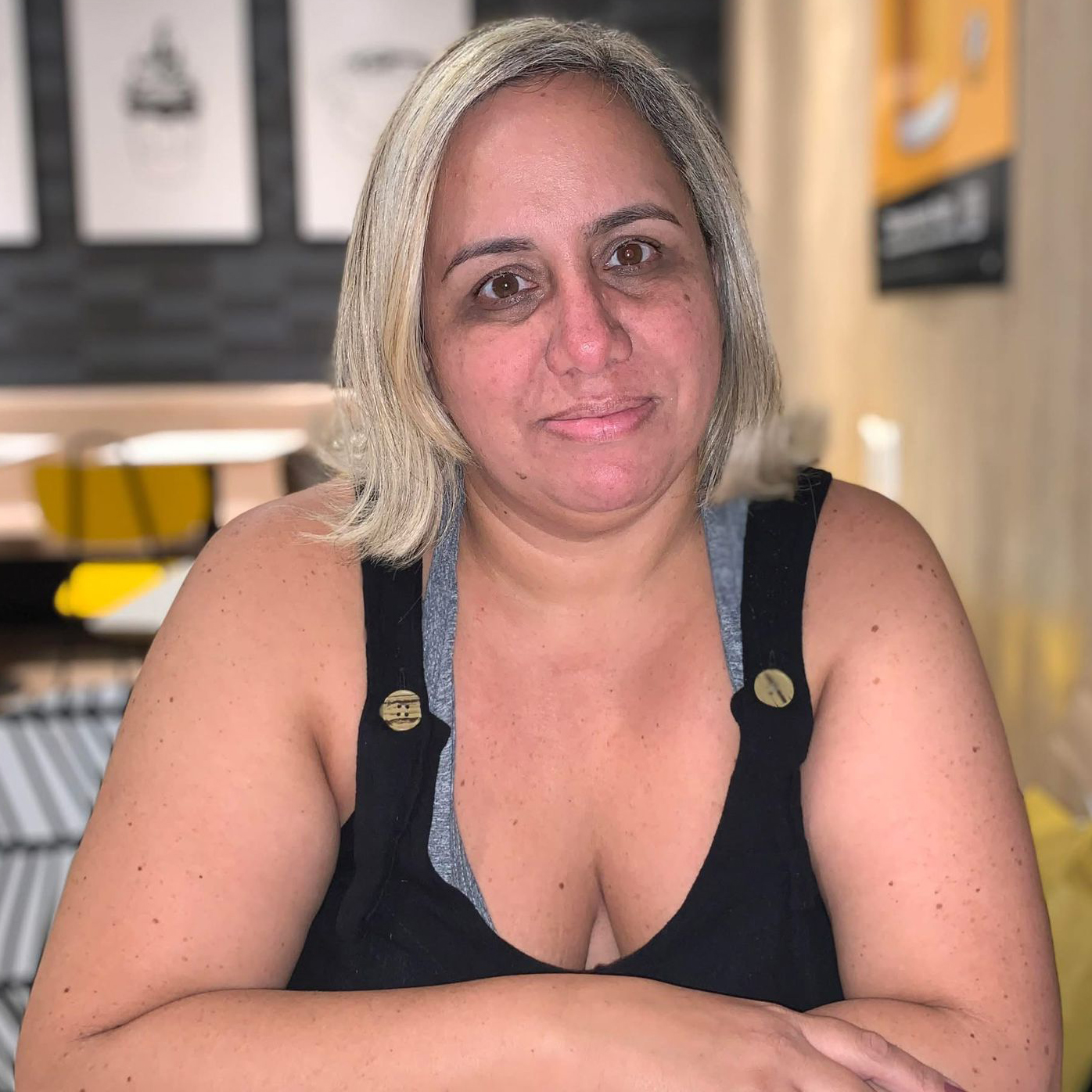 Kelly é uma mulher branca, de cabelos loiros, lisos, na altura dos ombros, repartidos levemente de lado. Tem sobrancelhas bem finas, olhos cor de mel e rosto arredondado. Usa uma blusa cinza por baixo de um macacão preto sem mangas, com botões dourados nas alças. Ela está sentada, com os braços apoiados e cruzados sobre a mesa. Ao fundo cadeiras e quadros pendurados na parede