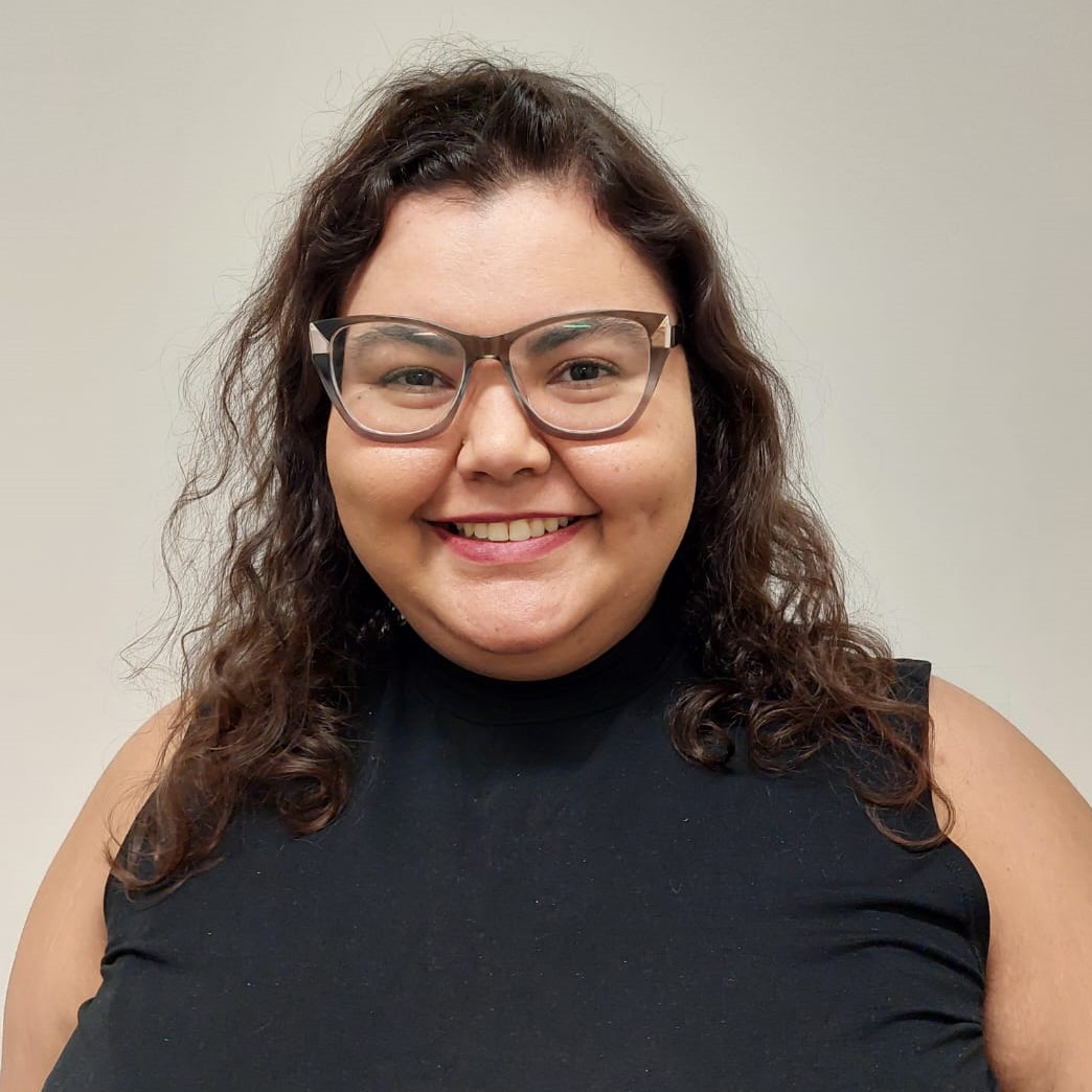 Descrição da imagem: Marina é uma mulher adulta, cisgênero, de pele clara, cabelos castanhos ondulados e usa óculos quadrados cinza. Ela sorri para a foto, na qual usa uma blusa preta sem mangas.