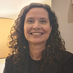 Roberta é uma mulher adulta, branca, olhos castanhos, com cabelos compridos e cacheados. Na foto está sorrindo, usa brincos e veste camisa e blazer pretos
