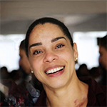 Solange é uma mulher de cabelos escuros, presos em um coque. Sorri para a foto e veste uma camisa marrom